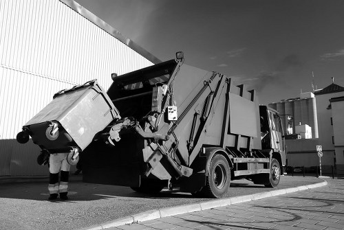 A commercial waste collection operation in progress with a large, modern refuse collection truck parked on a paved urban street near a high, corrugated metal boundary wall and adjacent to a tall white industrial building or warehouse. The truck is fitted with a hydraulic lifting mechanism at the rear, used for emptying large black wheelie bins, which are visible being lifted and tilted into the waste compartment. The bins appear to be made of durable plastic with textured surfaces, and are positioned in a manner indicating they are either being loaded or emptied. The scene is outdoors with natural daylight, and a person wearing work gloves and high-visibility trousers is seen handling a bin on the left side of the image. The setting has a tidy, functional appearance typical of commercial waste collection services in Acton, supporting the services provided by Commercial Waste Acton for rubbish removal in the local area.