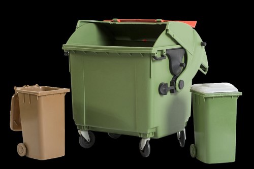 The image features three wheeled rubbish bins placed outdoors against a solid black background. The largest bin in the center is made of sturdy green plastic with a textured surface, a hinged lid in a matching green tone, and wheels at the base for mobility. To the left is a smaller, tan-colored bin, also plastic, with a smooth finish and two small wheels. On the right, a similar-sized green plastic bin with a ridged lid and wheels is positioned slightly behind the larger bin. The arrangement suggests a setup for waste collection or disposal, typical of commercial or residential rubbish removal services in Acton. The environment appears to be an outdoor area, possibly a driveway or yard, with no other visible contextual features. The lighting highlights the surfaces and details of each bin, emphasizing their textures and colors. The company name, Commercial Waste Acton, and the service focus on rubbish removal are subtly supported by the visual of multiple collection bins suitable for different types of waste, illustrating well-organized waste management equipment relevant to the local area.