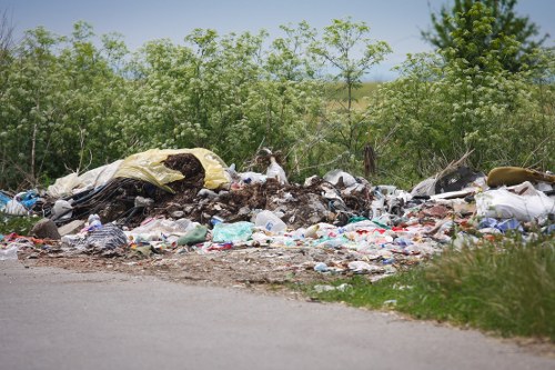 A large unmanaged pile of mixed rubbish is situated at the side of a paved road in an outdoor environment, with dense green trees and shrubs in the background. The debris includes various materials such as black and white plastic bags, shredded paper, broken household items, and discarded textiles. Some of the rubbish appears crumpled or torn, creating a heterogeneous landscape of waste with different textures and colours, predominantly in shades of white, black, and brown. The pile extends across a section of the roadside, partially encroaching onto the grassy verge, indicating the presence of illegal or unmanaged dumping. The setting suggests a rural or suburban area, potentially near Acton, which could require professional rubbish removal services like those offered by Commercial Waste Acton to prevent environmental contamination. The overall scene highlights the importance of proper waste disposal and environmental management, especially in areas prone to fly-tipping or illegal dumping.