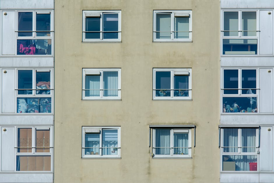 Photograph of a multi-story residential building with a brown brick facade, white walls, and large windows. Each of the four floors features a small balcony with dark metal railings, some with wooden
